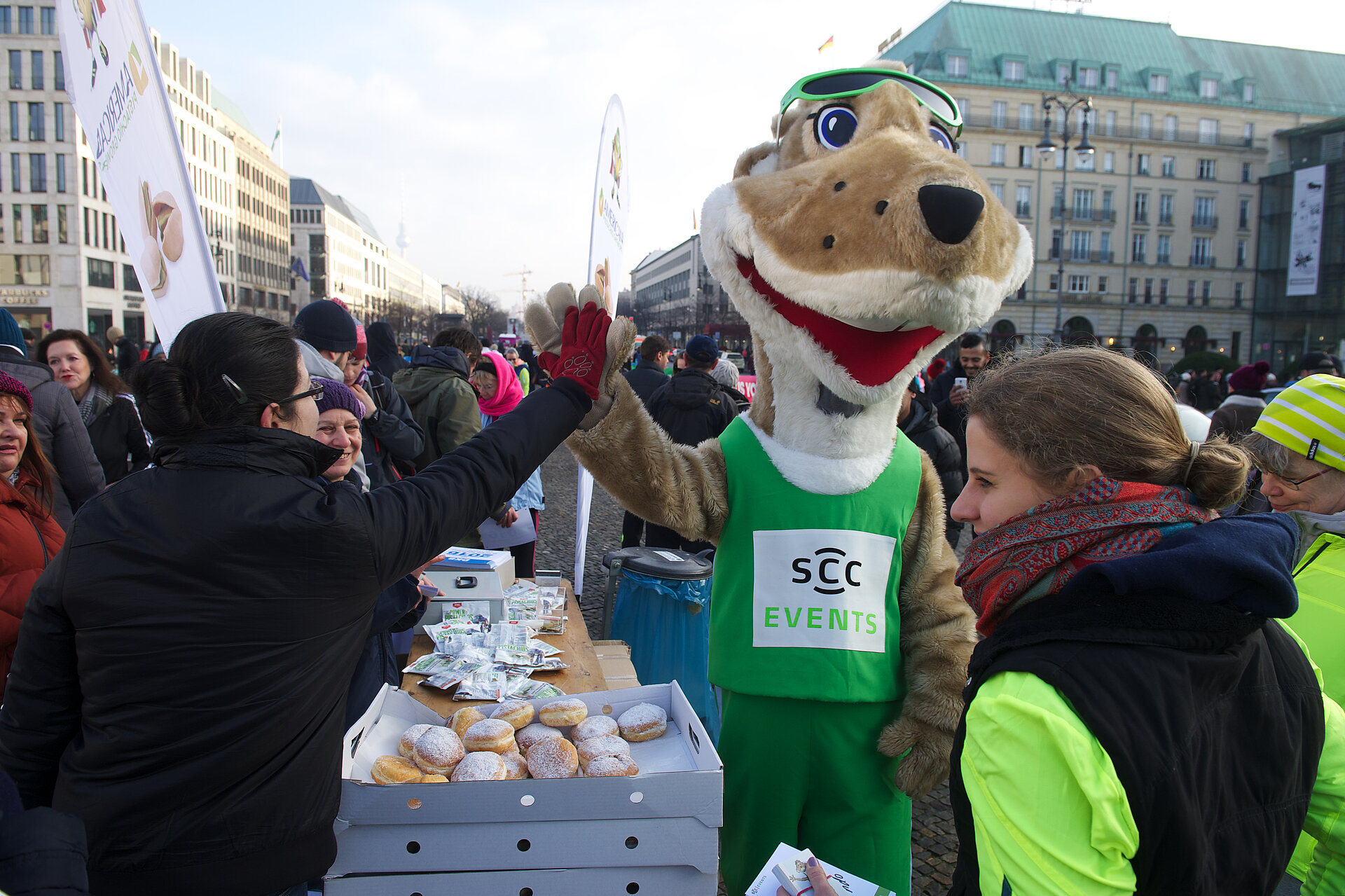 Berliner Neujahrslauf 2016: Frido klatscht Frau in schwarzer Jacke bei der Ausgabe der Pfannkuchen ab © SCC EVENTS / camera4