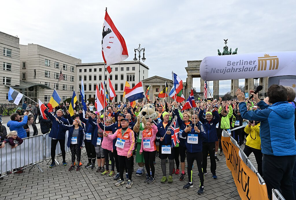 Berliner Neujahrslauf 2023: Spitze des Teilnehmerfeldes schwenkt Fahnen vor dem Start und wird von den Zuschauern bejubelt © SCC EVENTS / Petko Beier