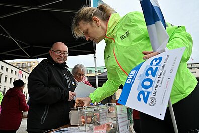 Berliner Neujahrslauf 2023: Weibliche Läuferin hält am Ausgabezelt Startnummer und Flagge in der Hand © SCC EVENTS/Petko Beier