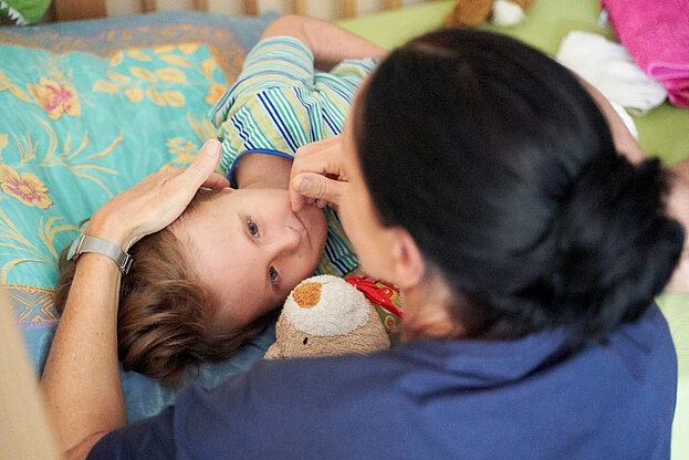 Weibliche Pflegekraft kümmert sich um eine Kind, welches im Bett liegt und einen Teddy im Arm hält © Björn Schulz Stiftung /Frank Nürnberger-Ole Oberländer