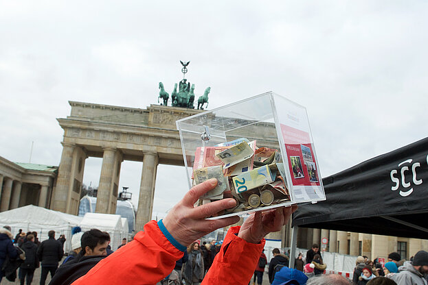 Berliner Neujahrslauf: Volunteer hält Spendenbox hoch, im Hintergrund das Brandenburger Tor © SCC EVENTS / camera4